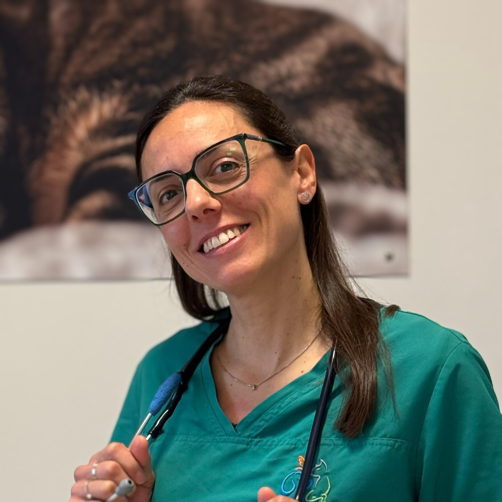 Alice Pierantoni, che indossa occhiali e un camice verde, sorride calorosamente mentre tiene uno stetoscopio appeso al collo. Lo sfondo sfocato aggiunge profondità alla scena.