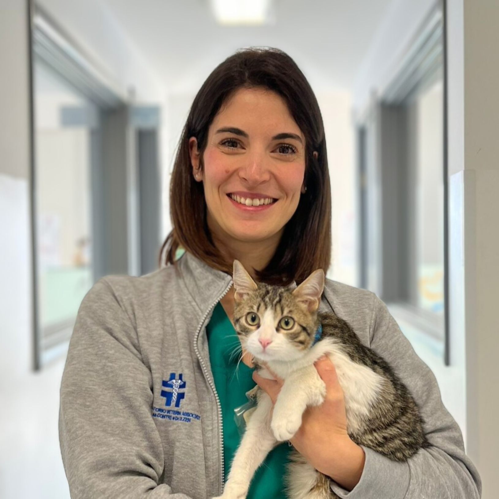 Claudia Bismondo, una donna con i capelli scuri e una giacca grigia, sorride mentre tiene in braccio un gatto soriano e uno bianco. Si trovano in un corridoio con porte su entrambi i lati, che suggeriscono una clinica o un ambiente veterinario.