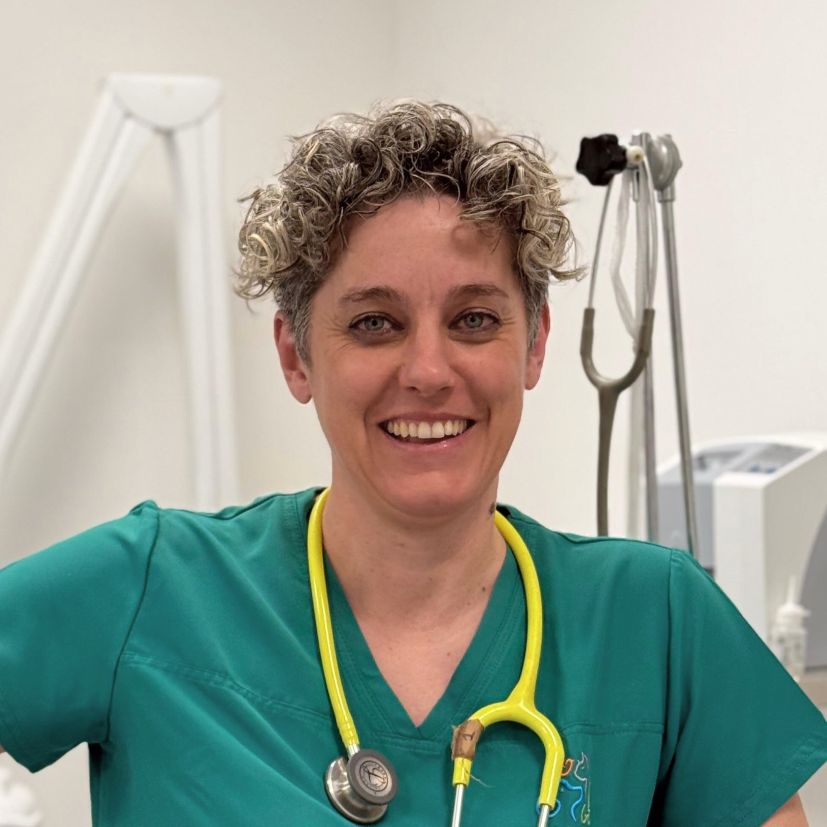 Lisa Pellattiero, in camice verde con uno stetoscopio al collo, sorride calorosamente in un ambiente medico. I suoi corti capelli ricci completano l'ambiente vivace, con le attrezzature mediche visibili sullo sfondo.