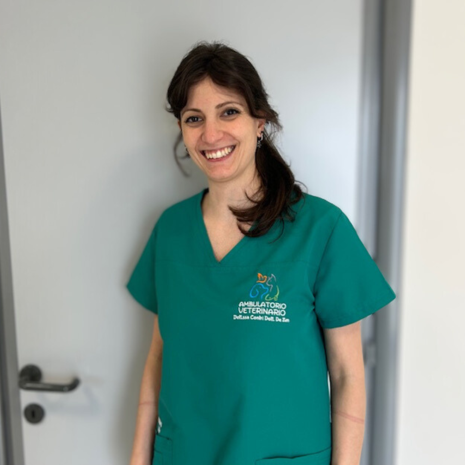 Miryam Modica, con i suoi lunghi capelli scuri e il suo sorriso caldo, indossa una camicetta verde ornata dal logo di una clinica veterinaria. Sta in piedi leggermente decentrata davanti a una porta grigia su uno sfondo semplice e chiaro.