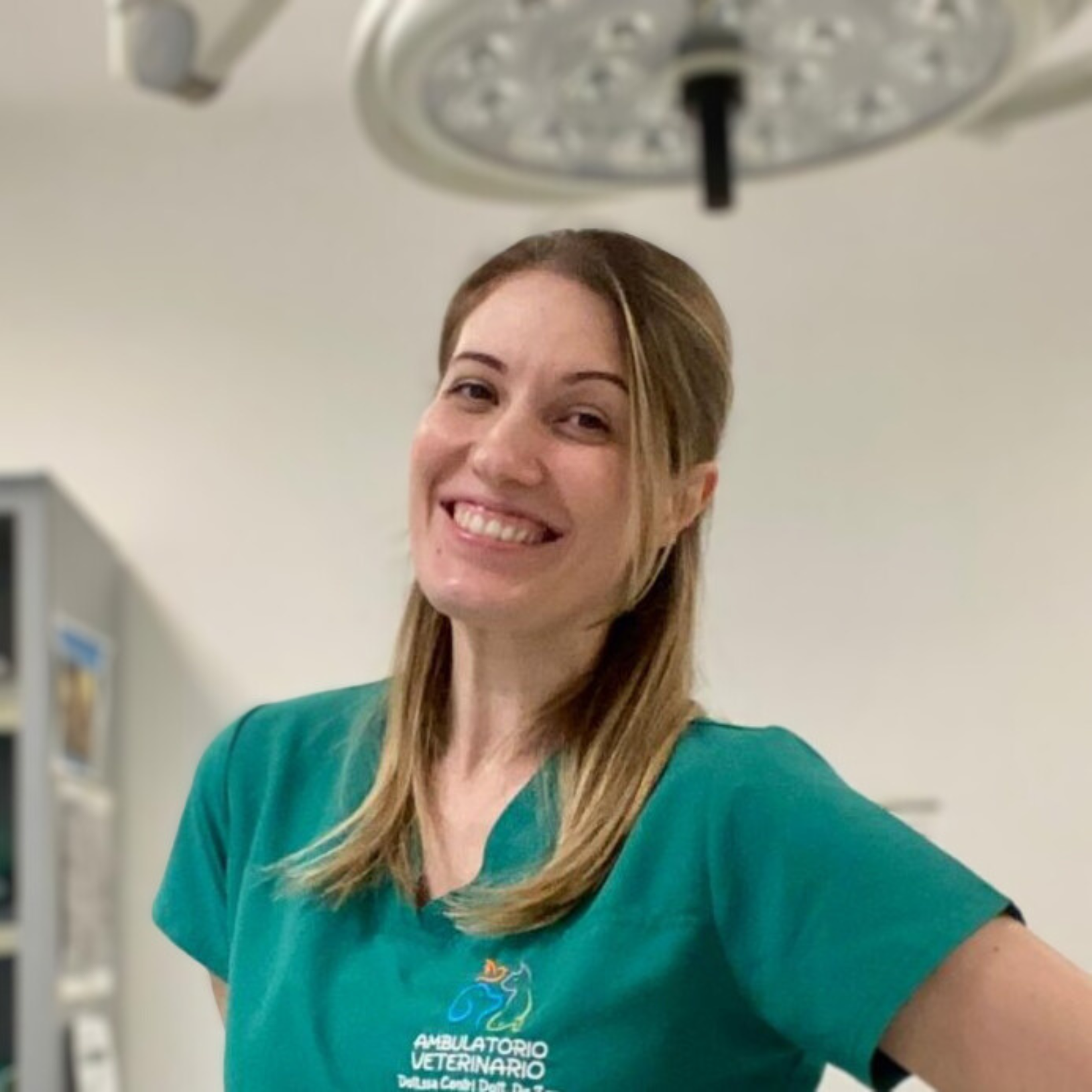 Una persona sorridente in camice verde, forse Stefania Dalla Riva, è in piedi in una stanza che ricorda una clinica veterinaria. Una luce dall'alto illumina la stanza e sullo sfondo si vede uno scaffale pieno di vari oggetti.