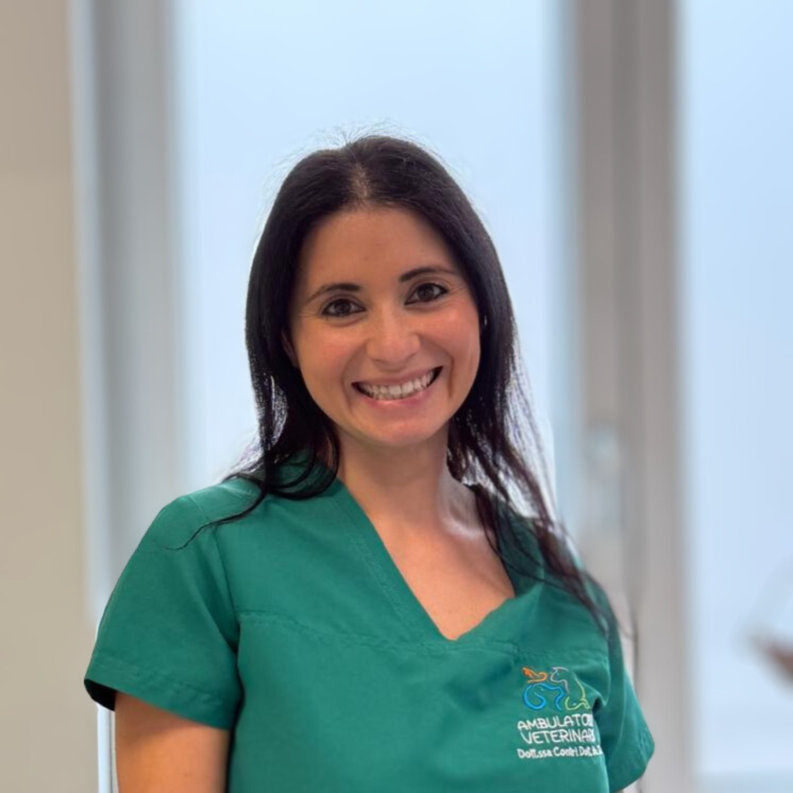 Teresa Auletta, una donna sorridente in camice verde, è in piedi all'interno, sullo sfondo di finestre leggermente sfocate.